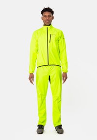 Veste imperméable jaune fluo avec un pantalon assorti, dotée d'un col haut, de poignets élastiques et d'une poche frontale zippée, associée à des chaussures foncées.