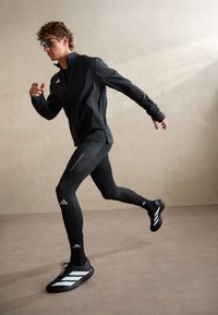 Uomo con abbigliamento sportivo nero e occhiali da sole che corre al chiuso contro una parete e pavimento beige uniforme.