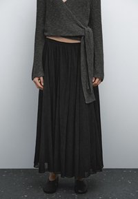 Personne portant un pull enveloppant gris foncé et une longue jupe plissée noire avec des chaussures noires à enfiler, le tout sur un fond uni.