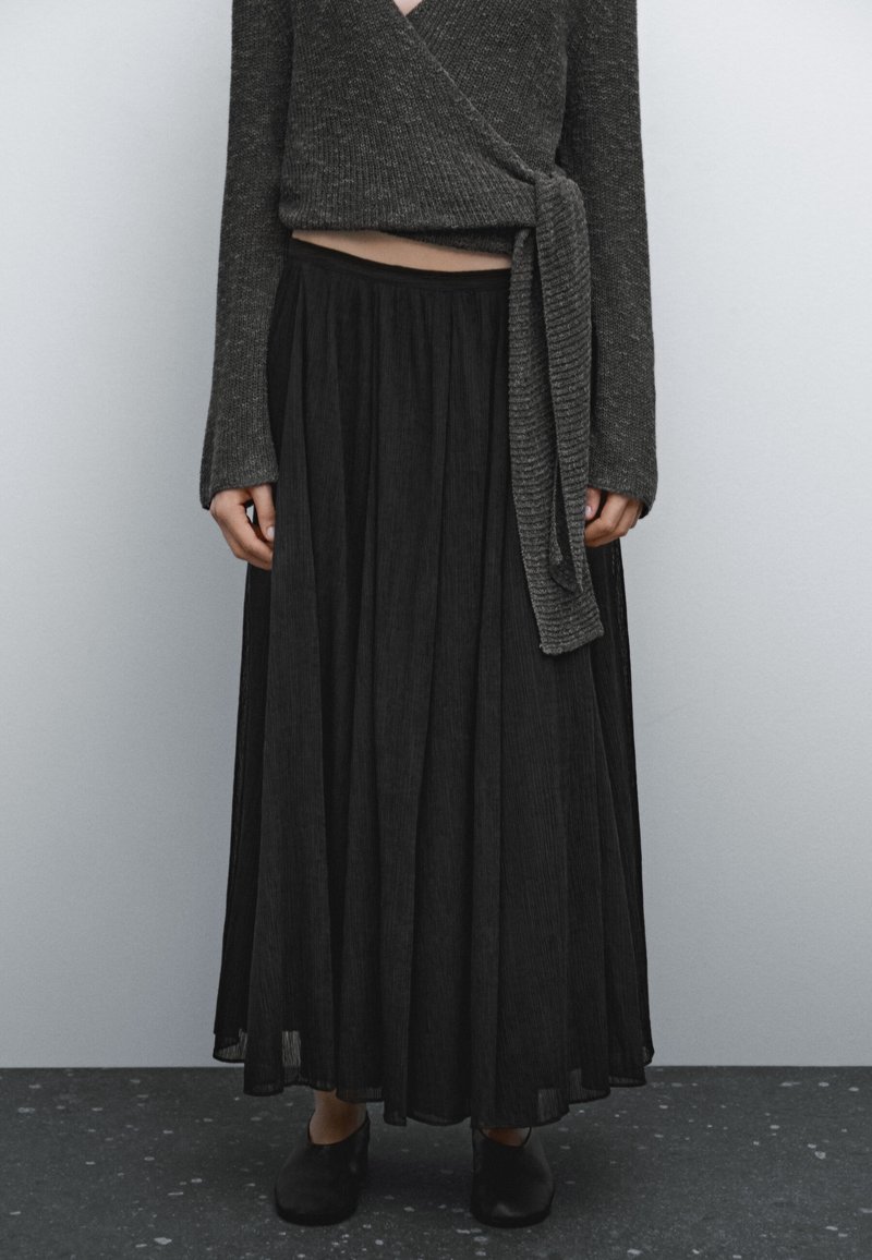 Personne portant un pull enveloppant gris foncé et une longue jupe plissée noire avec des chaussures noires à enfiler, le tout sur un fond uni.