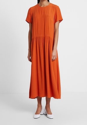 Kobieta w pomarańczowej sukience midi z krótkim rękawem i marszczoną talią oraz białych butach na słupku, stojąca na jednolitym tle.