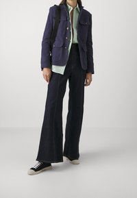 Chaqueta azul marino con cierres de botón y múltiples bolsillos, superpuesta a una camisa verde clara, combinada con jeans acampanados oscuros y zapatillas negras.