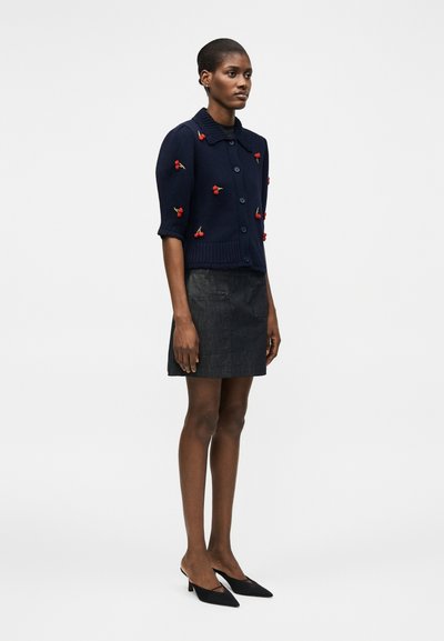 Γυναίκα που φοράει μια κοντομάνικη ζακέτα ναυτικού μπλε με κόκκινες εφαρμογές κερασιών, μια σκουρόχρωμη γκρι φούστα και μαύρες μυτερές γόβες, που στέκεται πλαγίως.