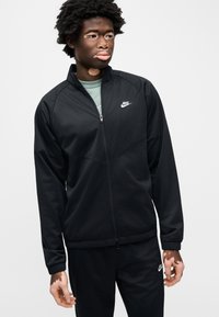 Μαύρο μπουφάν Nike με πλήρη φερμουάρ, ψηλό γιακά και ελαστικά μανσέτα. Ομαλό ύφασμα με απλό σχέδιο, λογότυπο στο αριστερό στήθος.