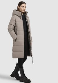 Vastag bézs színű hosszú kabát kapucnival, cipzáras zsebekkel és letisztult sziluettel; fekete fűzős katona cipőkkel párosítva.