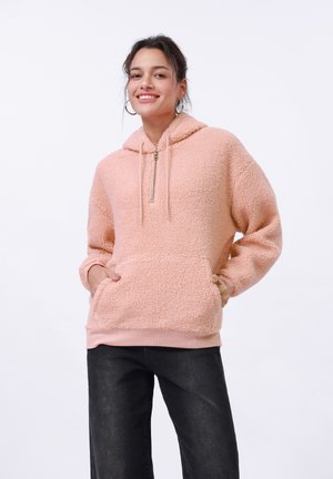 Light pink fleece hoodie with a zipper, kangaroo pocket, and soft texture; features a drawstring hood and ribbed hem. Paired with dark pants.