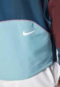 Chaqueta de Nike con un diseño bicolor en tonos de azul y marrón, logo blanco, textura suave y costuras rosas que contrastan.