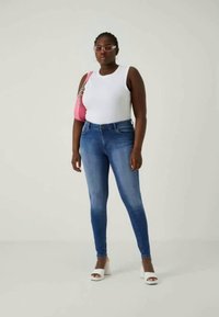 Jeans skinny de ganga azul com um design de cintura alta, combinados com uma blusa sem mangas branca e uma mala rosa. Modelo a usar sapatos brancos de salto.