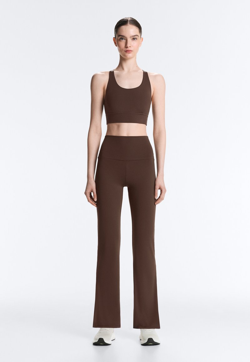 Kvinde stående frontalt iført brun ærmeløs croptop og høj taljede brune udvidede bukser med hvide sneakers på ensfarvet baggrund.