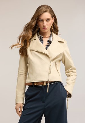 Donna con capelli mossi che indossa una giacca in pelle beige corta, camicia a righe, pantaloni blu navy, cintura marrone e orecchini pendenti dorati su sfondo neutro.