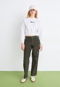 Sudadera gris recortada con el logo "obey", pantalones de pierna ancha en color verde oliva y zapatillas beige; el modelo lleva una gorra beige con un diseño simple.