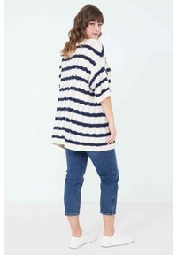 Dicker Strickcardigan in Creme mit Marineblau gestreift, übergroßer Schnitt, kurze Ärmel und gerippter Saum. Kombiniert mit blauen, kurzen Jeans und weißen Sneakers.
