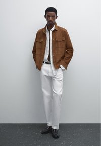 Giacca in suede marrone con due tasche sul petto, camicia bianca, pantaloni bianchi e mocassini neri. Sfondo semplice, modello in piedi con le braccia rilassate.