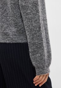 Grijze gebreide trui van textuurstof, met een loszittende mouw en geribde manchetten. Draagt over marineblauwe streepjaspantalons.