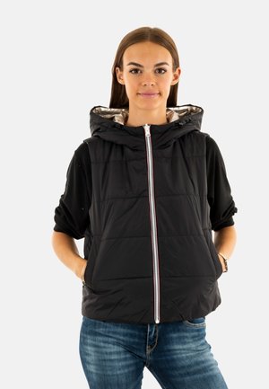 Gilet imbottito nero con chiusura frontale a zip, cappuccio regolabile e sottile bordo bianco e rosso. Texture trapuntata, abbinata a una maglietta nera a maniche lunghe e jeans blu.