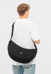 Czarna torba na ramię o zakrzywionym kształcie, wykonana z tkaniny. Posiada regulowany pasek oraz małą łatę z logo. Gładka tekstura bez widocznych wzorów.