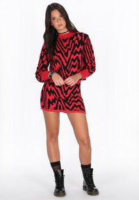 Rochie-pulover tricotată, cu un model îndrăzneț în zigzag roșu și negru, cu mâneci lungi, manșete ribbate și o margine deasupra coapsei. Se poartă cu cizme de luptă negre.