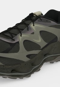 Gros plan d’une chaussure de sport grise et noire avec une maille texturée, des superpositions ondulées, des lacets noirs et une semelle épaisse tachetée.