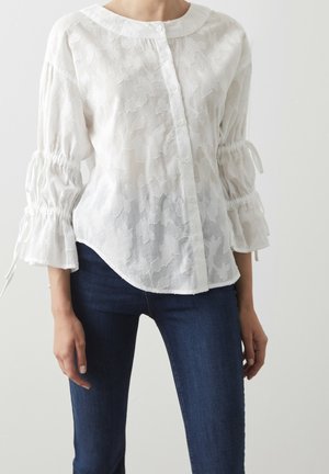 Femme portant un chemisier blanc à texture florale avec des manches froncées et un jean bleu foncé ajusté, debout devant un fond uni.