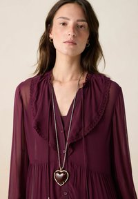 Robe bordeaux en tissu transparent superposé, ornée de dentelle et de boutons. Comprend un long collier argenté avec un pendentif en forme de cœur.
