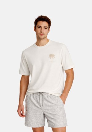 Ung mand iført en hvid t-shirt med et palmetræsgrafik og lysegrå leopardprintshorts, stående med den ene hånd i lommen.