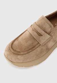 Mocassin en suede marron clair avec un bout légèrement surélevé et arrondi, et une semelle plate et rembourrée. Présente une sangle décorative sur le dessus.