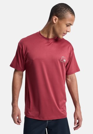 Mężczyzna w prostym czerwonym t-shircie z krótkim rękawem i małym białym logo na lewej piersi, patrzący w dół w prawo.