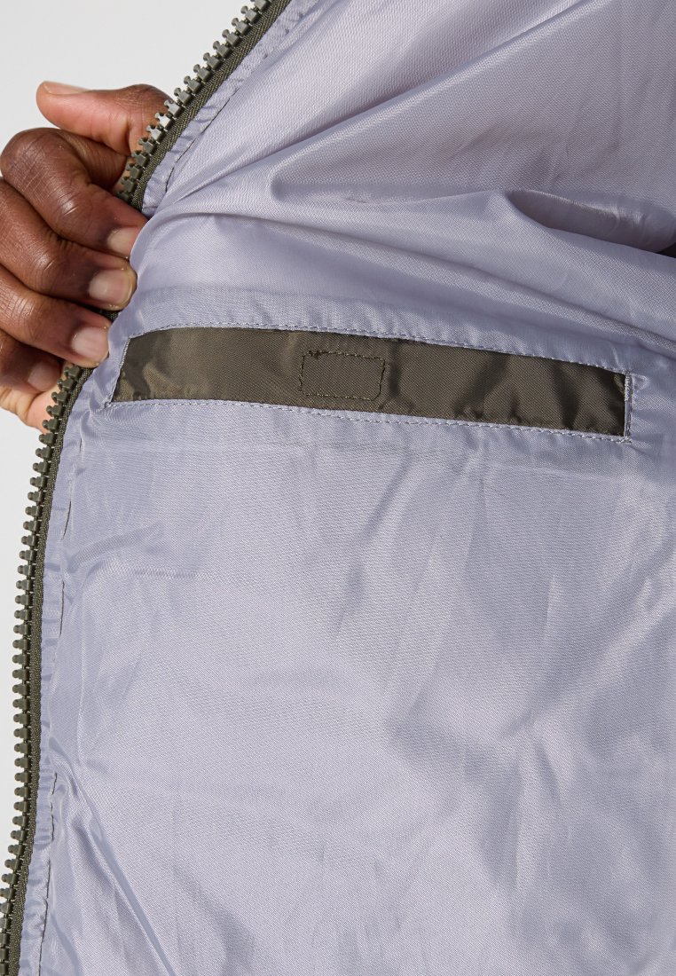 Chaqueta ligera con cremallera, con un interior de forro suave en lavanda pálido y un bolsillo oculto bordeado de tejido verde oliva oscuro.