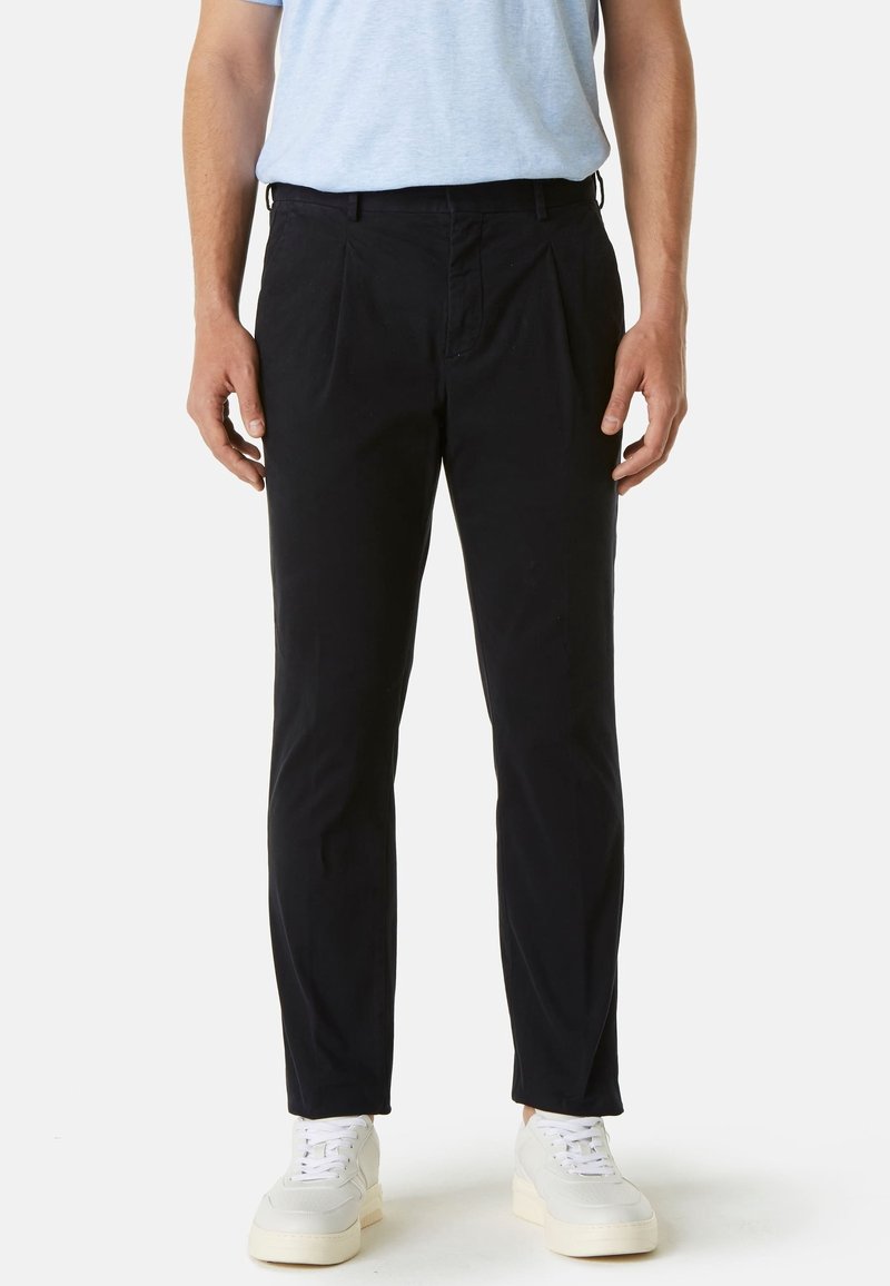 Muž v černých slim-fit kalhotách, světle modrém tričku a bílých teniskách, stojící proti jednoduchému světlému pozadí.