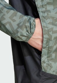 Chaqueta ligera con puños elásticos, que presenta una manga con un patrón verde y un cuerpo negro. El patrón incluye rayas verticales y formas.