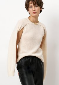 Licht beige wollen sweater met een ontspannen pasvorm, brede lange mouwen en een open schouderontwerp, gecombineerd met zwarte leren broeken.