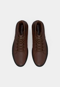 Brązowe skórzane sneakersy za kostkę z okrągłym noskiem, czarną gumową podeszwą i sznurowadłami; wewnątrz buta znajduje się oznakowanie marki Converse.