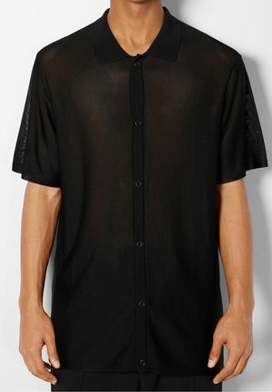 Mężczyzna ubrany w czarną półprzezroczystą koszulę z krótkim rękawem, kołnierzykiem i guzikami, stojący na jednolitym tle.