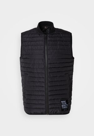 Musta puffervesti, joka on valmistettu tikatusta kankaasta, jossa on korkea kaulus, vetoketjukiinnitys, sivutaskut ja logopatchi vasemmassa alareunassa.