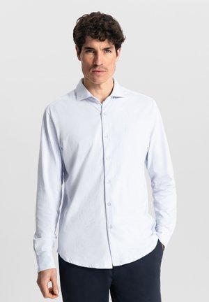 Camisa de manga larga azul claro con cuello clásico y cierre de botones, que presenta una textura suave y un sutil detalle de logo en el pecho.
