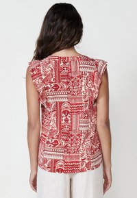 Top senza maniche con motivo rosso, caratterizzato da un mix di design intricati, con dettagli arricciati sulle spalle e uno sfondo beige chiaro.