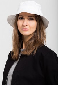 White bucket hat with a wide brim, smooth fabric, worn by a person with medium-length brown hair, paired with a black jacket and grey top.