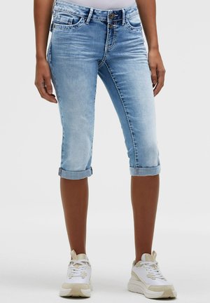 Frau trägt hellblaue hochgekrempelte Denim-Caprihosen und weiße Sneakers und steht vor einem einfarbigen Hintergrund.