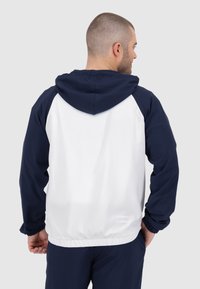 Giacca con cappuccio navy e bianca, caratterizzata da una texture liscia, polsini elasticizzati e una vita arricciata. Presenta un design a blocchi di colore solido.