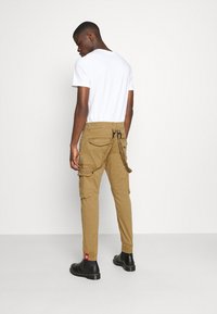 Cargo-bukser i khaki med multiple lommer, en snøre bagpå og struktureret stof. Sammen med en ensfarvet hvid T-shirt og sorte støvler.