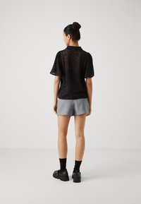 Svart blonder skjorte med korte ermer og krage, kombinert med lysegrå shorts og svarte klumpete sko. Glatt tekstur med blomsterdesign.