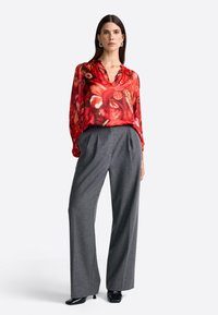 Kvinde stående iført en rød blomstretd blouse med lange ærmer, grå vide bukser, sorte stiletter og sølvøreringe.