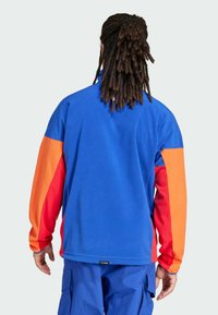 Fleece pulover u plavoj boji s narančastim i crvenim rukavima. Ima visoki ovratnik, opušteni kroj i logotip na donjem rubu. Glatka tekstura.