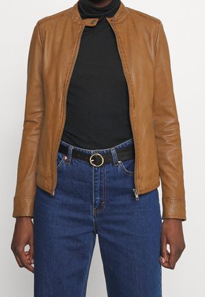 Osoba nosící béžovou koženou bundu přes černý rolák, zastrčený do vysoké modré džínové sukně s černým pásem s kulatou zlatou sponou.