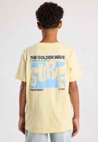 Lichtgele katoenen t-shirt met korte mouwen, met opvallende grafische tekst in blauw en zwart op de achterkant: "DE GOUDE Golf SURF PACIFIC BEACH 1989".
