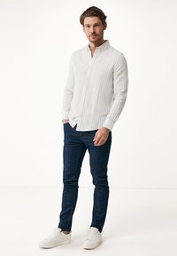Hombre de pie con las manos en los bolsillos, vistiendo una camisa blanca de botones con rayas claras, jeans azul oscuro y zapatillas blancas, frente a un fondo blanco.