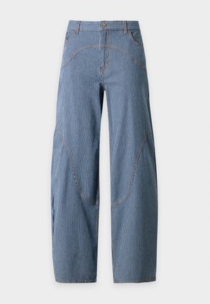 Femme aux longs cheveux blonds portant une chemise blanche à manches courtes avec col, un large pantalon bleu rayé et des ballerines noires pointues, debout avec une main dans la poche.