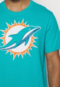 Buste masculin portant un t-shirt turquoise avec un grand logo dauphin devant un design rayons de soleil orange.