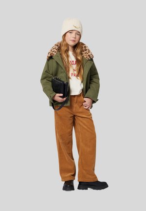 Veste verte avec col en imprimé léopard, chemise blanche avec texte rouge, pantalon en velours côtelé marron, sac à main noir, béret crème. Bottes noires épaisses.