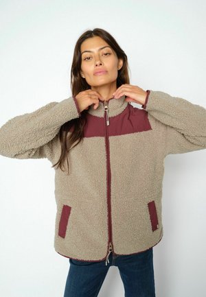 Kvinde står mod en ensfarvet baggrund iført en beige og bordeaux fleecejakke med lynlås og blå jeans, justerer kraven.